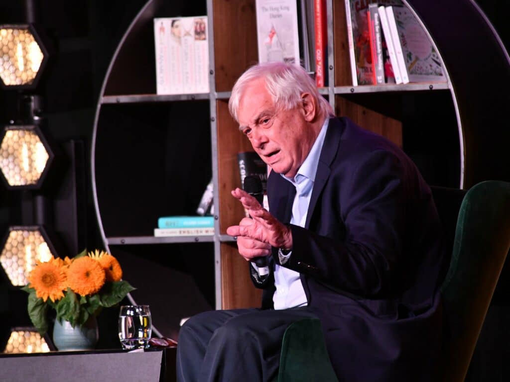 Chris Patten giving a talk on stage in front of a bookshelf