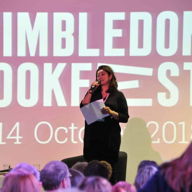 Fiona Razvi on stage in front of an audience with a microphone, in front of a Wimbledon Bookfest advert