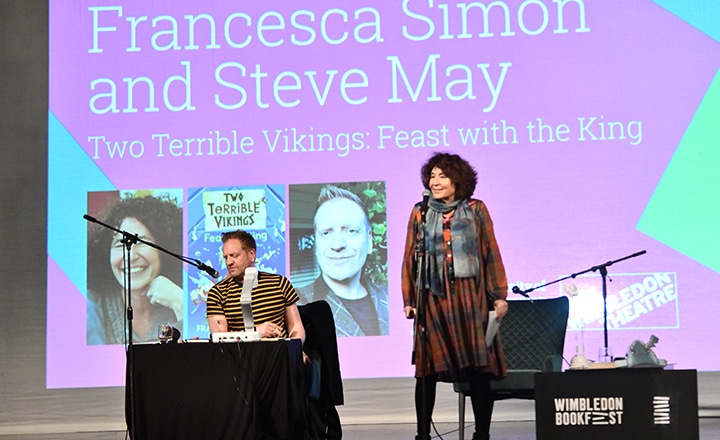 Author Francesca Simon stood on stage talking into a microphone with Author Steve May sat down on stage. Their book Two Terrible Vikings : Feast with the King on a screen behind them