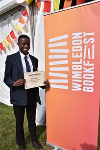 A proud competition winner holding a certificate stood by a Wimbledon Bookfest banner