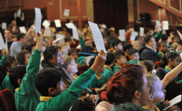 A Group of school children in an audience, all holding up pieces of paper